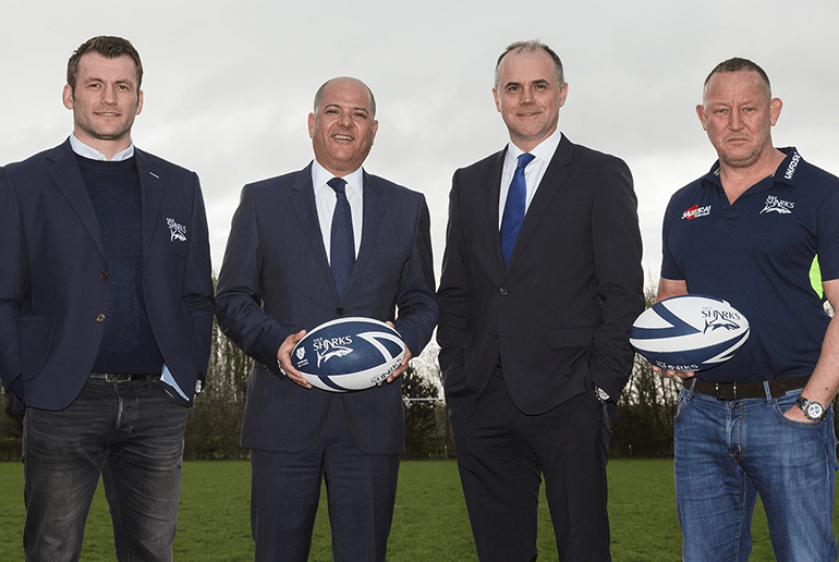 four men standing next to each other holding sale shark rugby balls