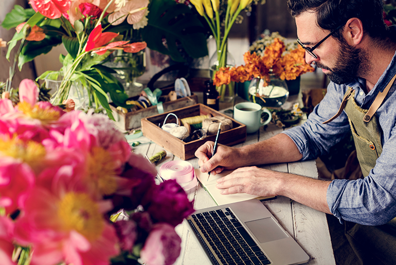 man wearing an apron surrounded by flowers sat at a table with a laptop writing in a notepad