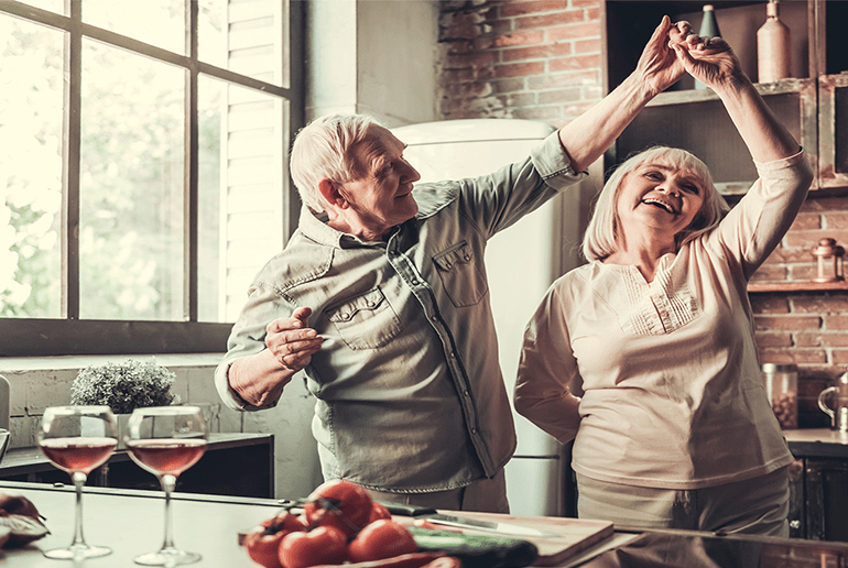 older couple dancing in a kitchen whilst cooking with two glasses on wine on the side