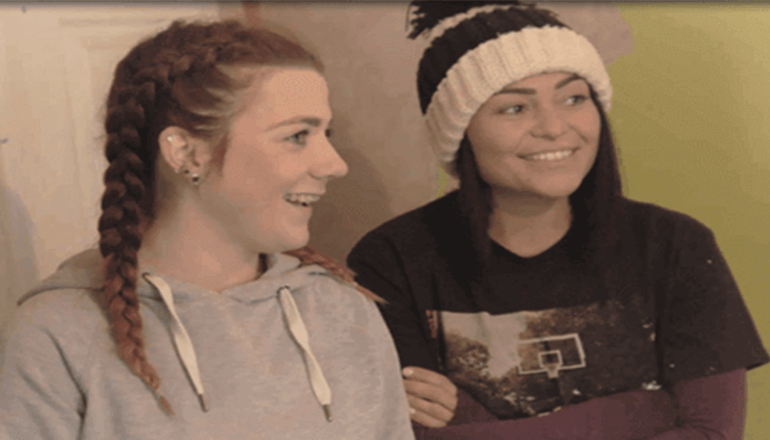 two girls stood next to each other smiling one with plaited hair and one wearing a hat