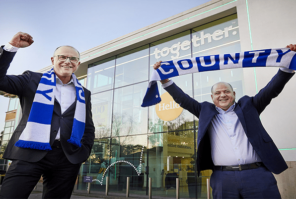 two men in suits outside a building holding stockport country scarfs - l