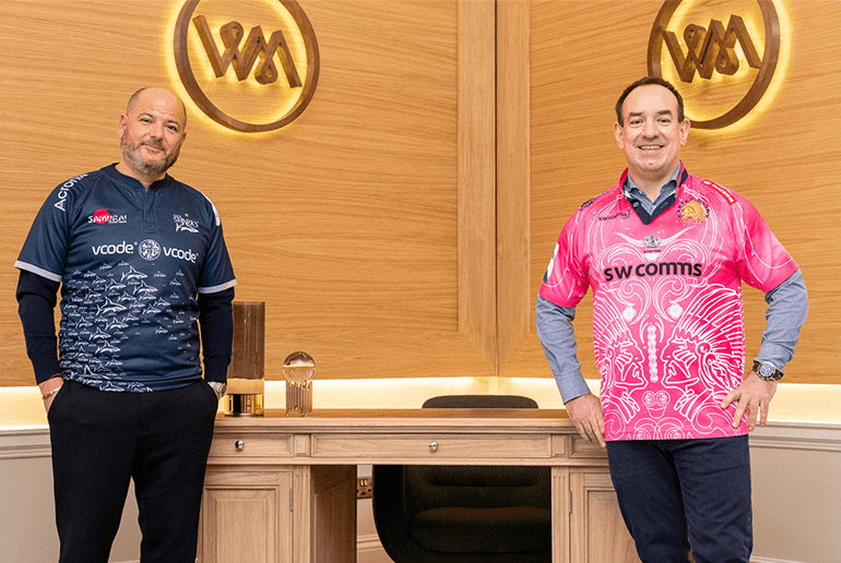 Two men smiling with a sale sharks t-shirts on stood in front of winslade park desk - l