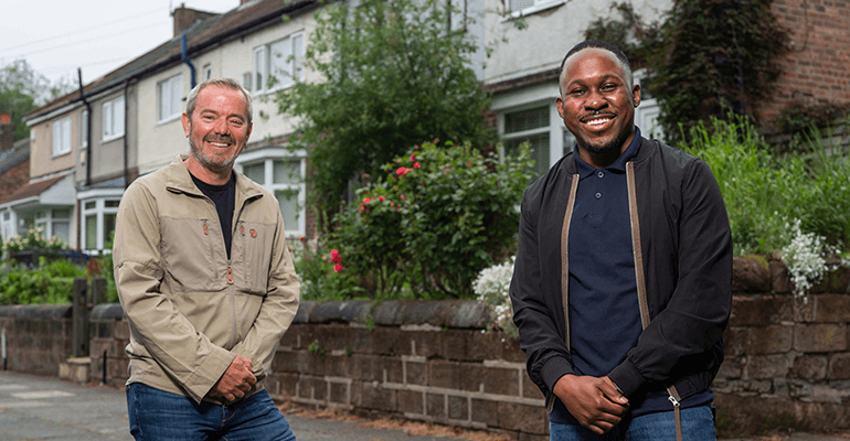 Two men standing near each other smiling in front of a building.