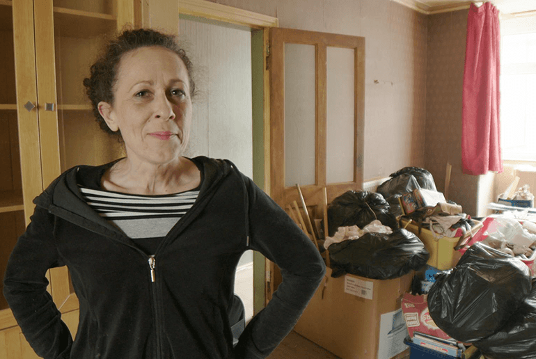 woman in black zipped cardigan stood in a living room with a pile of full bin bags behind her