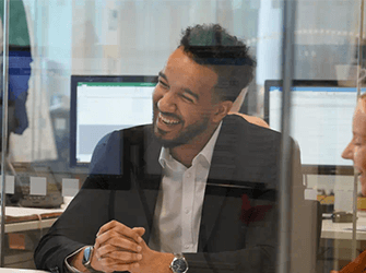 A close up of a man sat at a desk in a black suit laughing.