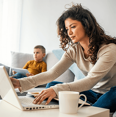 A woman busy on her laptop with her child watching TV in the background.