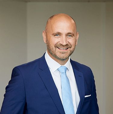 A bald man with a stubble smiling wearing a navy blue suit with a white shirt and a light blue tie.