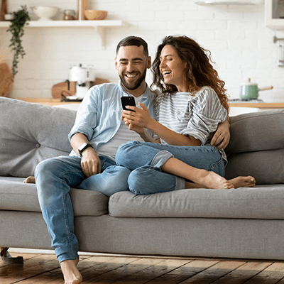 happy couple sat on a grey couch looking at a mobile phone