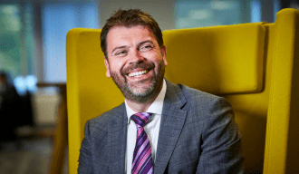 Man smiling wearing grey suit with white shirt and purple tie sat in a yellow chair