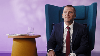 A man with black hair sat on a blue chair looking into the distance.